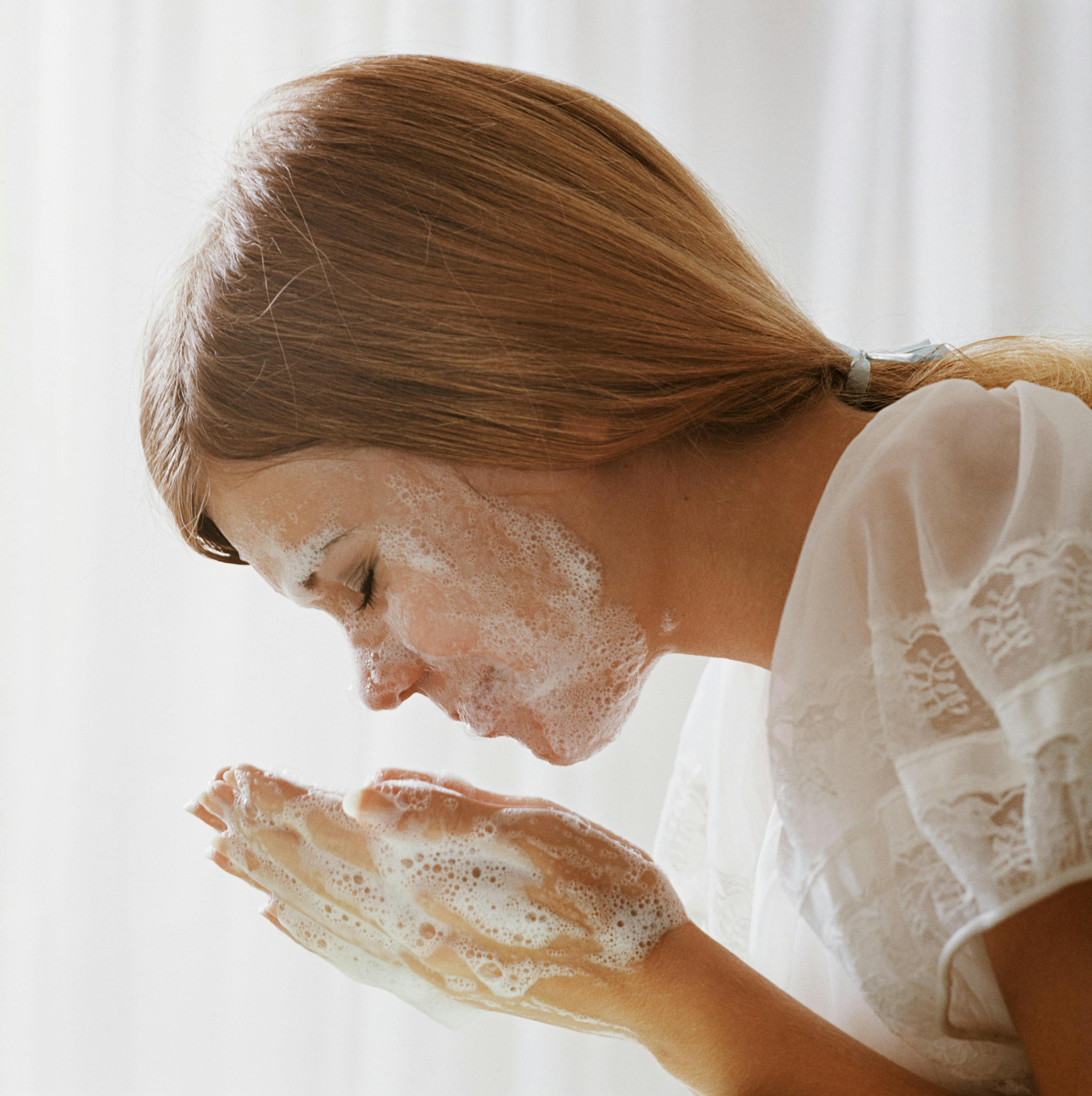Một người phụ nữ với mái tóc xoăn bồng bềnh đang rửa mặt trong chiếc áo phông trắng. Cô ấy đang giơ tay lên mặt, tay và mặt cô ấy nổi đầy bọt vì sữa rửa mặt của cô ấy có bọt.